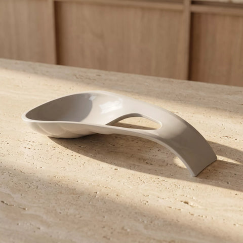 A modern, curved ceramic dish with a central cutout, resting on a textured beige stone surface with warm sunlight and a wooden cabinet in the background.