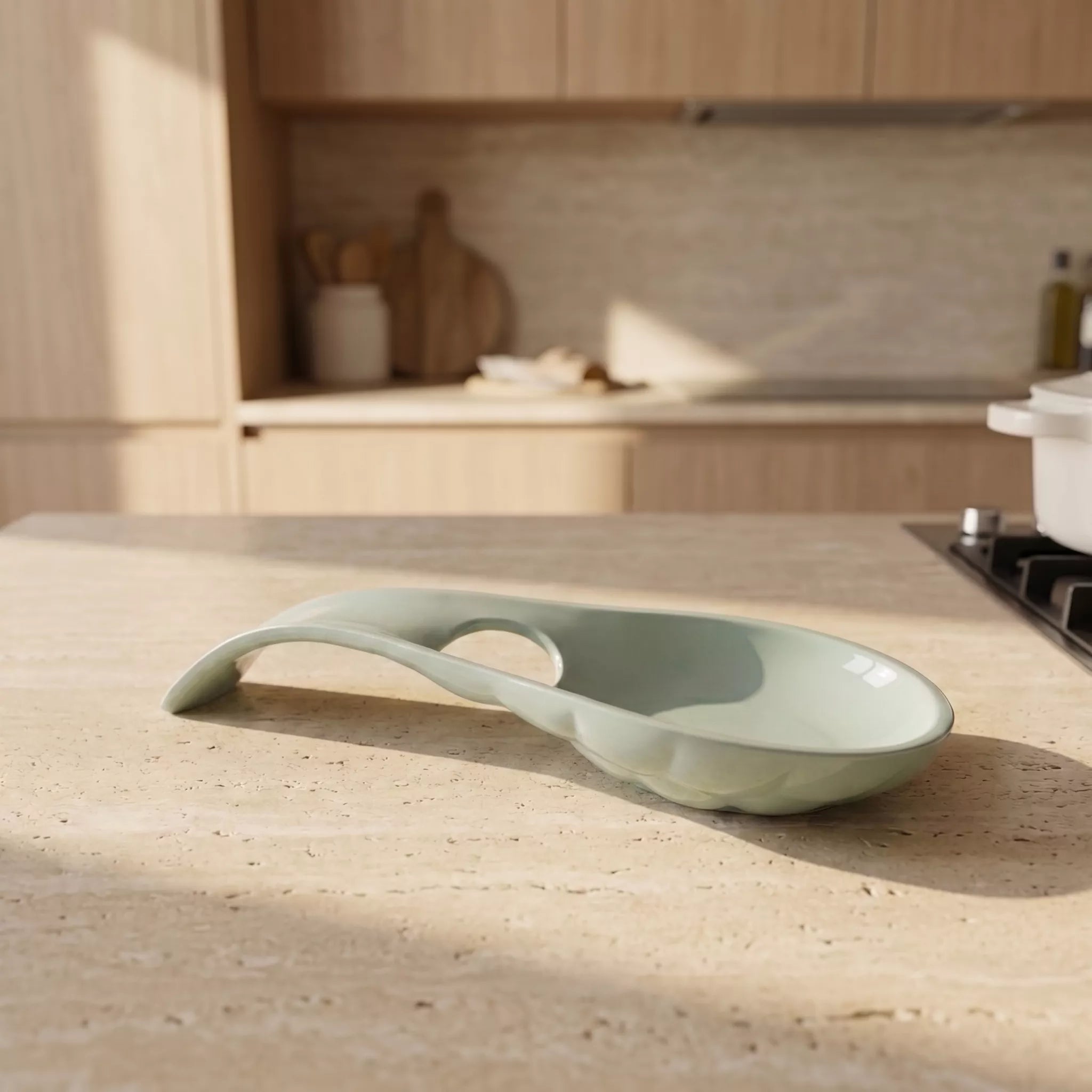 A light sage green ceramic spoon rest with a loop handle, resting on a beige stone countertop in a softly blurred kitchen setting.