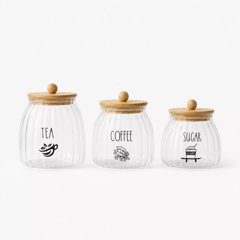 Three ribbed clear glass canisters with light wood lids and black printed labels for TEA, COFFEE, and SUGAR, arranged on a white background.