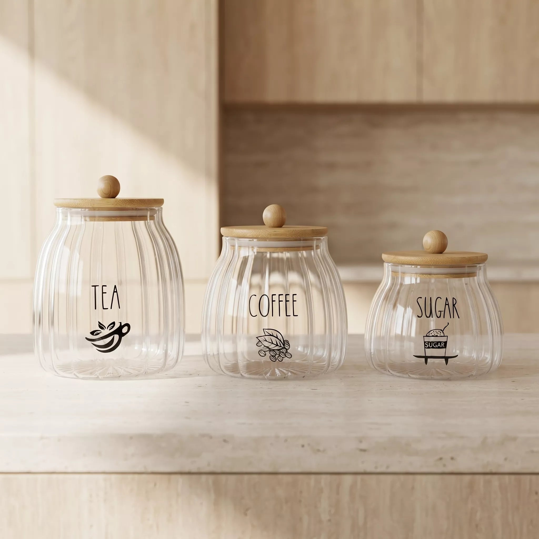 Three ribbed clear glass canisters with light wood lids and round knobs, labeled TEA, COFFEE, and SUGAR, arranged on a light wood countertop in a softly lit kitchen setting.