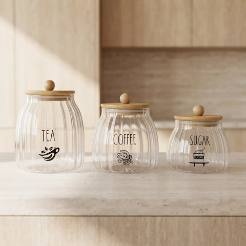 Three ribbed clear glass canisters with light wood lids and round knobs, labeled TEA, COFFEE, and SUGAR, arranged on a light wood countertop in a softly lit kitchen setting.